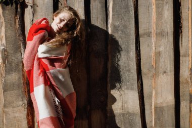 Genç kadın dışarıda güneş ışığında ahşap bir duvara karşı kırmızı ve beyaz bir ekose sarılmış ayakta