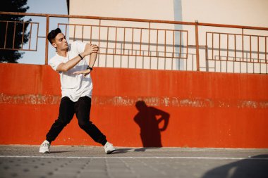 Kot giyinmiş ve beyaz t-shirt şık genç adam güneşli bir günde boyalı beton duvara karşı sokakta brakedance dans ediyor