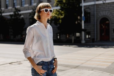 Beyaz gömlek ve kot giyinmiş beyaz güneş gözlüğü şık genç kadın yaz güneşli bir gün şehir sokakta ayakta