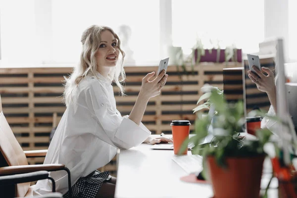 Ung, vakker kvinne i hvit skjorte jobber med en telefon ved skrivebordet med en bærbar PC i et moderne, åpent romkontor. – stockfoto