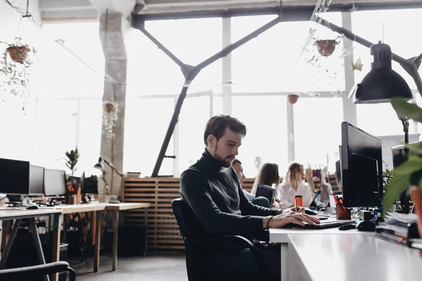 Fyren i uformelle kontorklær jobber med datamaskinen ved siden av kollegene i det moderne lyskontoret. – stockfoto