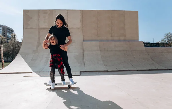 젊은 아버지는 화창한 날에 스케이트 공원에서 스케이트 보드를 타는 어린 아들을 돕습니다. — 스톡 사진
