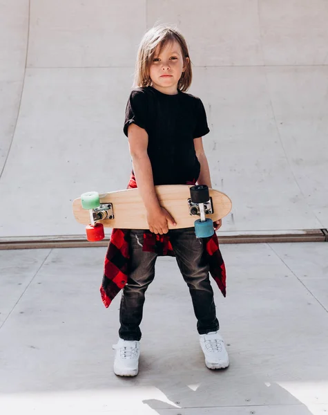 De jongen gekleed in de casual kleding met skateboard in zijn hand staat in een skatepark naast de glijbaan op de zonnige dag buiten — Stockfoto