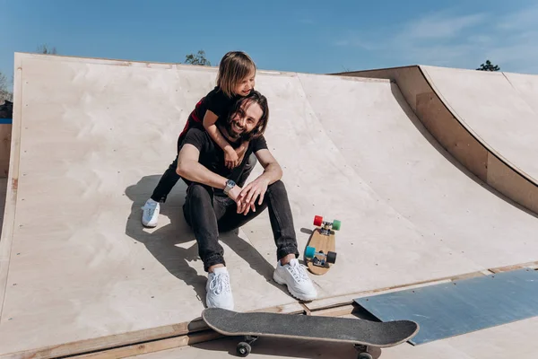 Happy young father and his son dressed in the stylish casual clothes are sitting in an embrace together on the slide next to the skateboards in a skate park at the sunny warm day