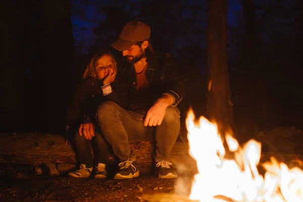 Feliz padre y su hijo se calientan por el fuego sentado en un abrazo en los troncos en una caminata en el bosque por la noche . — Foto de Stock