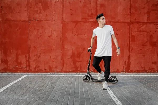 Pemuda berpakaian jins dan t-shirt berdiri dengan skuter terhadap dinding beton dicat pada hari musim panas di kota . — Stok Foto