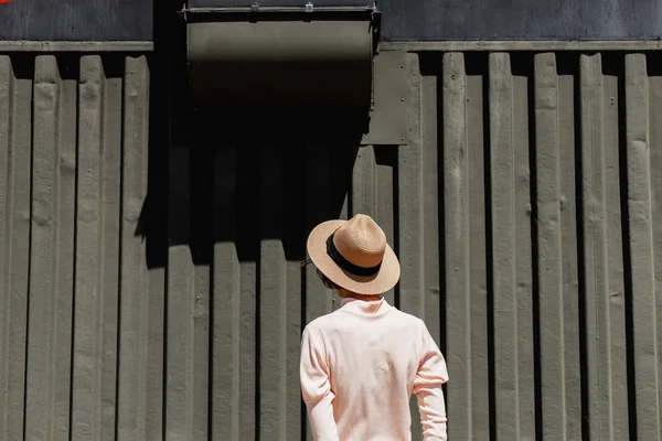 Stijlvol meisje in een zacht roze jurk en stro hoed staan naast een donkere metalen hek op een zonnige dag — Stockfoto