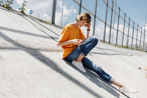 노란 티셔츠와 청바지에 재미 소녀는 외부 화창한 날에 유리 울타리 옆에 콘크리트 슬래브에 앉아있다 — 스톡 사진
