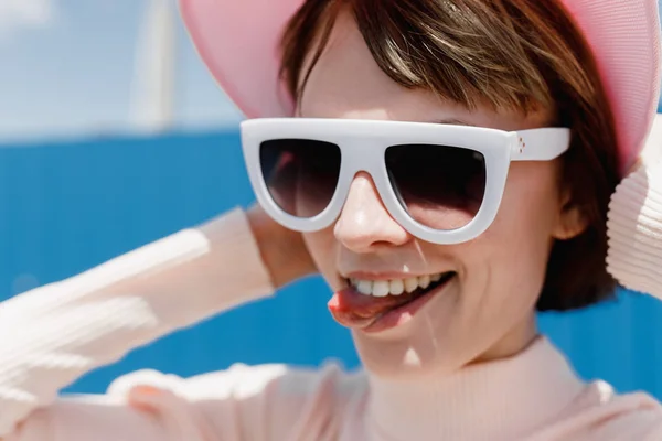 Menina engraçada em óculos de sol brancos e viseira rosa mostra sua língua em um dia ensolarado fora — Fotografia de Stock