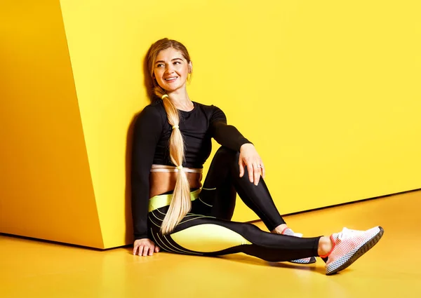 The athletic girl with long blond hair dressed in a stylish sportswear is sitting on the yellow floor next to the yellow wall.