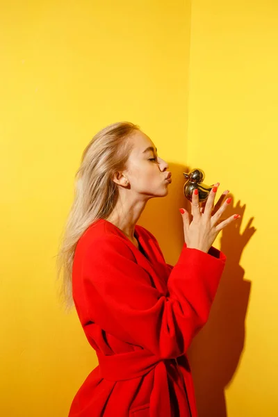 Dívčí módní blogovací figurína oblečená ve stylovém červeném kabátě má na sobě zlatou malou kachničku v dlaních na pozadí žlutých stěn v pořadu pokoje — Stock fotografie