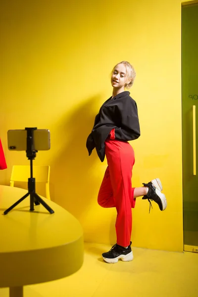La joven bloguera de moda vestida con pantalones rojos y chaqueta negra se toma una selfie en el teléfono inteligente de pie sobre un soporte en la mesa de la habitación con paredes y muebles amarillos — Foto de Stock