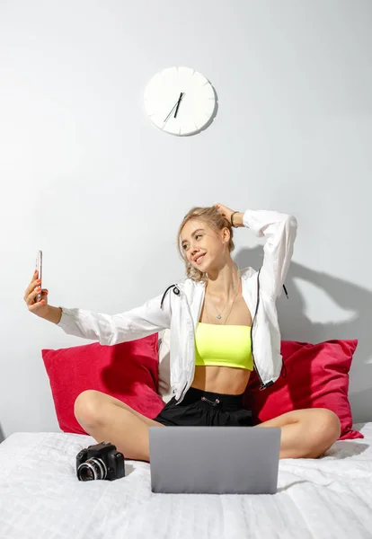 Посміхаючись молода дівчина блогер одягнений в модний одяг бере селфі на свій смартфон для її блог, сидячи на ліжку з ноутбуком в кімнаті — стокове фото