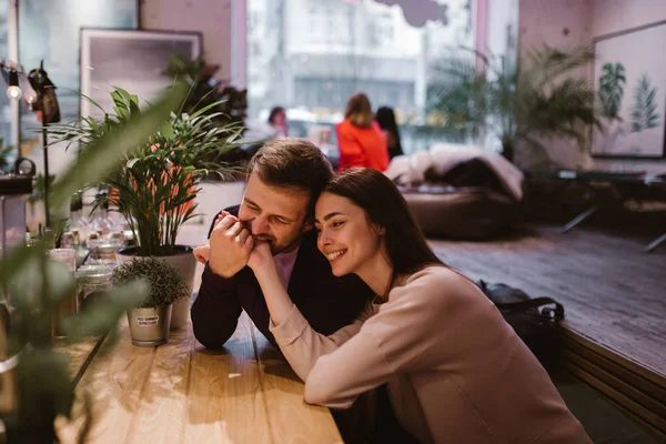 Cara carinhoso feliz segura e morde sua mão namoradas sentado na mesa no café e olha para ela . — Fotografia de Stock