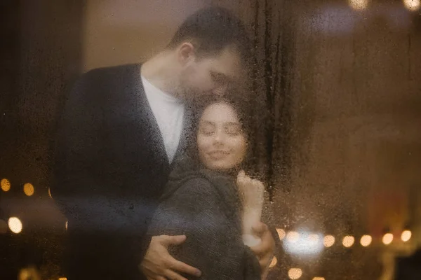 Cara amorosa abraça e beija sua namorada de pé atrás de uma janela molhada com luzes. Casal romântico — Fotografia de Stock