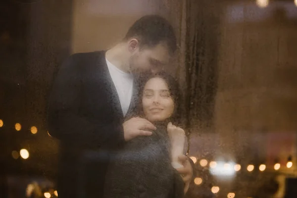 Cara amorosa abraça e beija sua namorada de pé atrás de uma janela molhada com luzes. Casal romântico — Fotografia de Stock