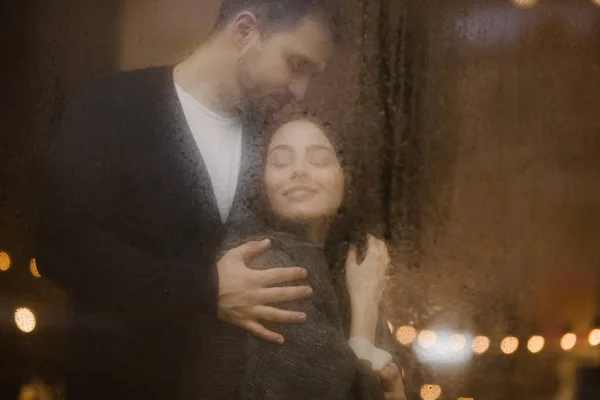 Feliz cara amoroso abraça sua namorada de pé atrás de uma janela molhada com luzes — Fotografia de Stock
