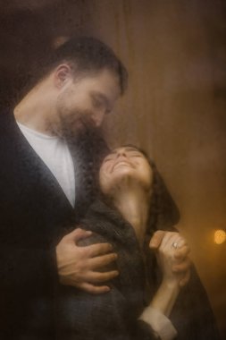 Romantik sevgi dolu adam ışıkları ile ıslak bir pencerenin arkasında duran onun mutlu kız arkadaşı kucaklar.