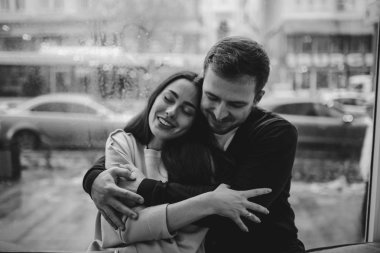 Loving adam rahat bir kafede pencere üzerinde oturan onun güzel mutlu kız arkadaşı kucaklar. Siyah beyaz fotoğraf