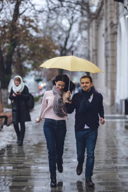 Güzel çift, adam ve rahat giysiler giymiş kız arkadaşı yağmurda sokakta şemsiye altında çalışan.