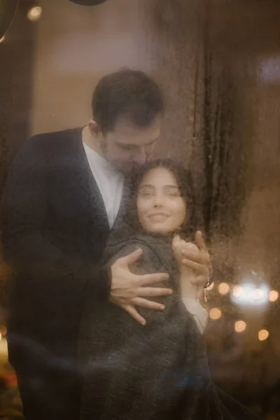 Romantic loving guy hugs his happy girlfriend standing behind a wet window with lights.