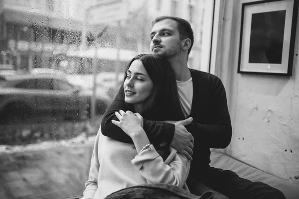 Romantic couple. Loving guy hugs his beautiful girlfriend sitting on the windowsill in a cozy cafe.