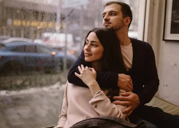 Una pareja romántica. chico cariñoso abraza a su hermosa novia sentada en el alféizar de la ventana en un acogedor café . —  Fotos de Stock