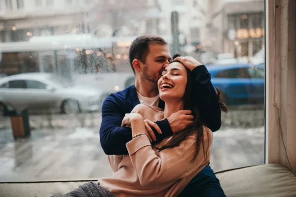 Liebevoller Kerl umarmt seine schöne, glückliche Freundin, die auf der Fensterbank in einem gemütlichen Café sitzt. — Stockfoto