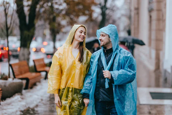 Cara e garota amorosa nas capas de chuva amarelas e azuis caminham na rua na chuva — Fotografia de Stock