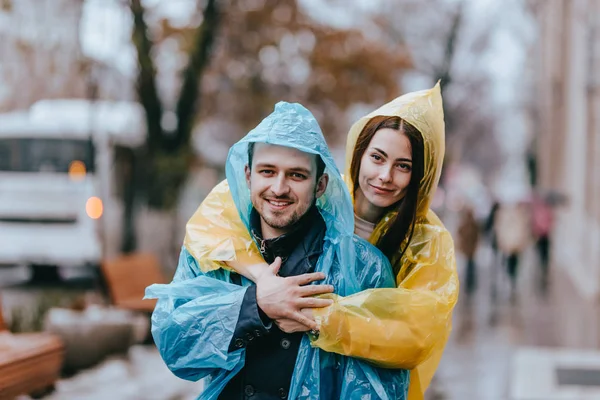 Feliz casal amoroso cara e sua namorada vestida com capas de chuva estão abraçando na rua na chuva — Fotografia de Stock