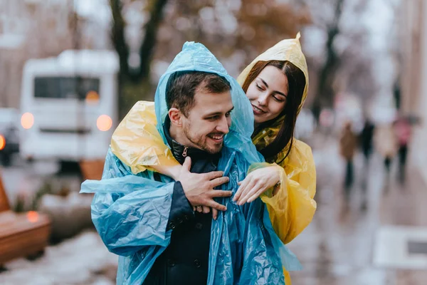 Feliz casal amoroso cara e sua namorada vestida com capas de chuva estão abraçando na rua na chuva — Fotografia de Stock