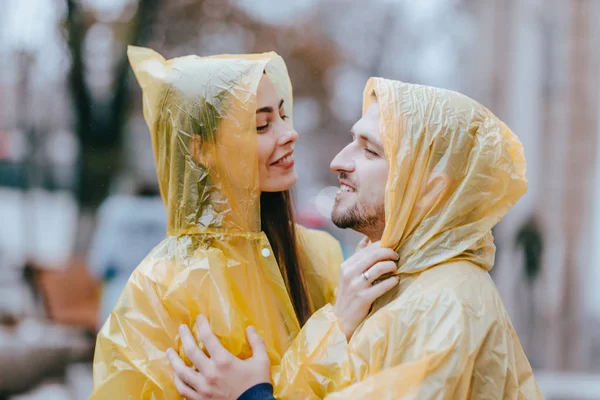 Feliz casal amoroso, cara e sua namorada vestida com capas de chuva amarelas estão abraçando na rua na chuva — Fotografia de Stock