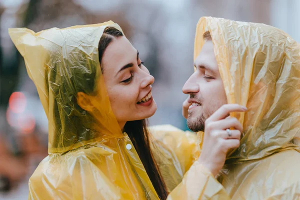 Casal amoroso, cara e sua namorada vestida com capas de chuva amarelas estão abraçando na rua na chuva — Fotografia de Stock