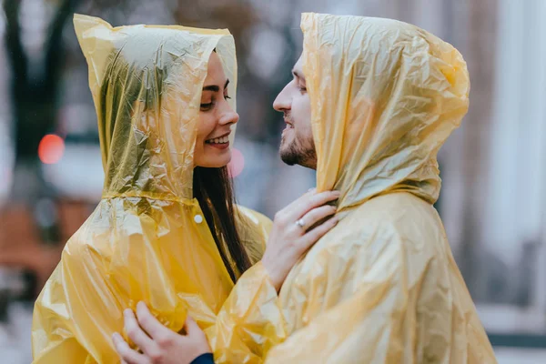 Casal amoroso, cara e sua namorada vestida com capas de chuva amarelas estão abraçando na rua na chuva — Fotografia de Stock