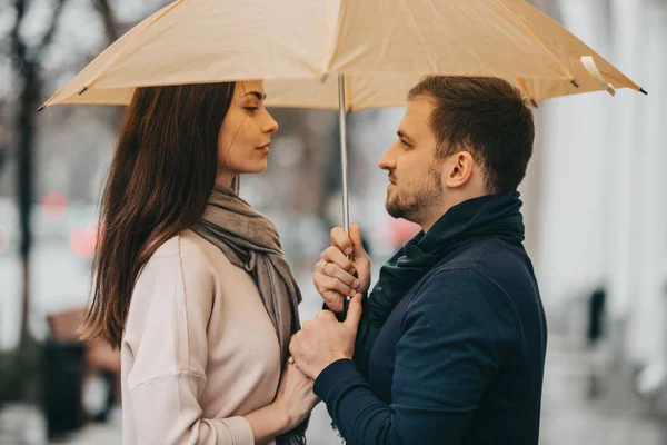 Vackra paret, killen och hans flickvän klädd i casual kläder står under paraplyet och titta på varandra på gatan i regnet — Stockfoto