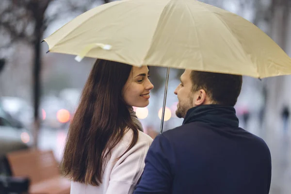 Vackra paret, killen och hans flickvän klädd i casual kläder står under paraplyet och titta på varandra på gatan i regnet — Stockfoto