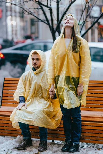 Guy and his girlfriend dressed in yellow raincoats standing near the bench on the street in the rain