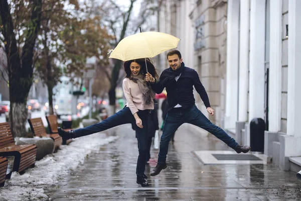 Mutlu çift, adam ve rahat giysiler giymiş kız arkadaşı yağmurda sokakta şemsiye altında atlıyorlar. — Stok fotoğraf