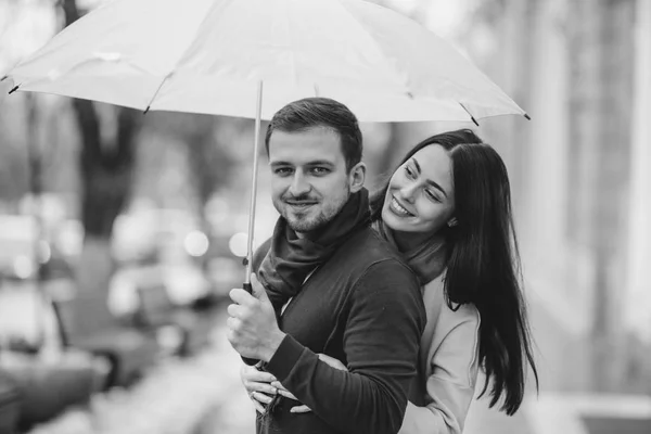 Casal romântico feliz, cara e sua namorada vestida com roupas casuais estão abraçando sob o guarda-chuva e olhar um para o outro na rua na chuva. Foto em preto e branco — Fotografia de Stock