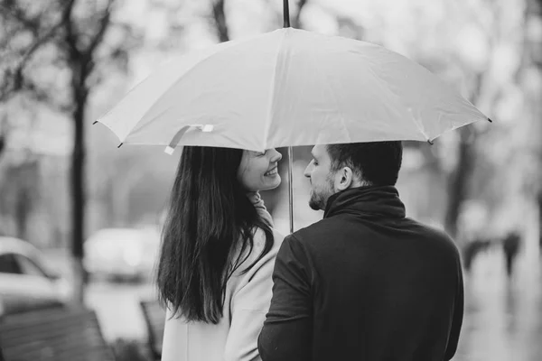 Feliz casal bonito, cara e sua namorada vestida com roupas casuais ficar sob o guarda-chuva e olhar um para o outro na rua na chuva. Foto em preto e branco — Fotografia de Stock