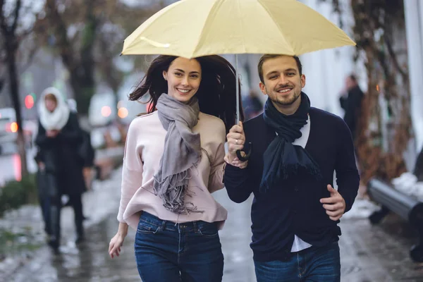 Beautiful couple, guy and his girlfriend dressed in casual clothes are running under the umbrella on the street in the rain. — Stock Photo, Image