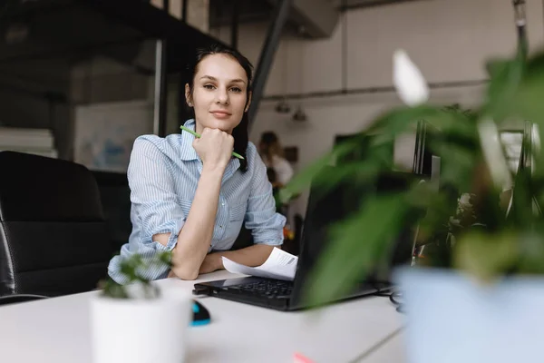 Young woman dressed in office style clothes is working at the laptop sitting at desk in the light modern office.