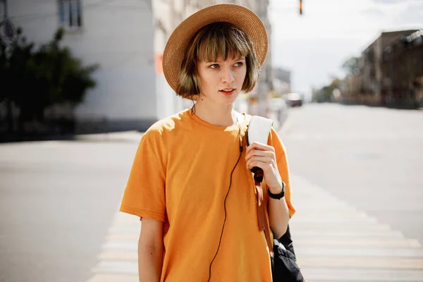 Молода дівчина з навушниками в жовтій футболці і солом'яний капелюх гуляє з рюкзаком вздовж міської вулиці в літній сонячний день — стокове фото