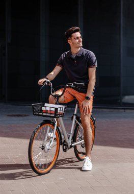 Şık adam moda rahat giysiler giymiş gri bir binanın arka planına karşı sokakta bisiklet sürmek