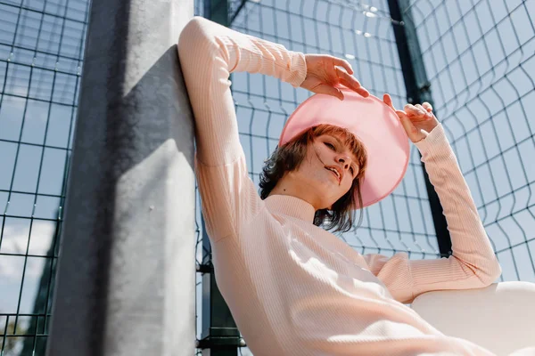 Hafif pembe elbise ve pembe vizör bir yaz güneşli bir günde spor alanında kafes çit arkasında bir sandalyede oturan şık kız