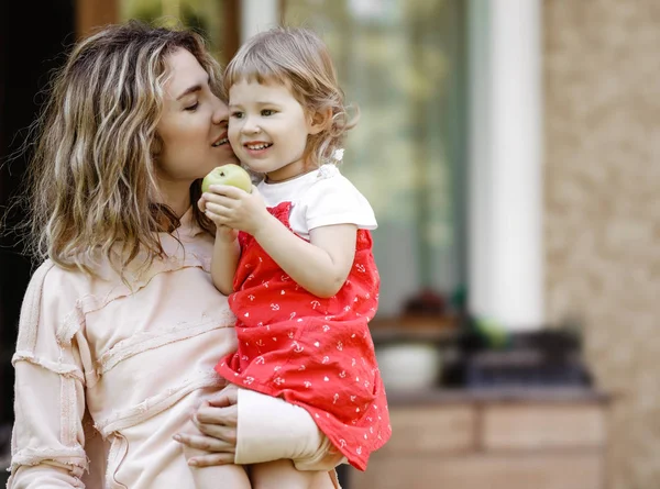 Junge Mutter hält ihre kleine Tochter auf den Händen und küsst ihre süße Wange an einem warmen Sommertag im Garten — Stockfoto