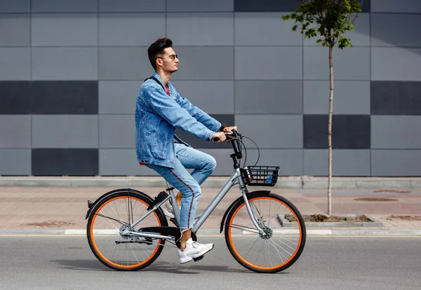 Pria bergaya berpakaian jins trendi mengendarai sepeda di jalan dengan latar belakang bangunan abu-abu — Stok Foto