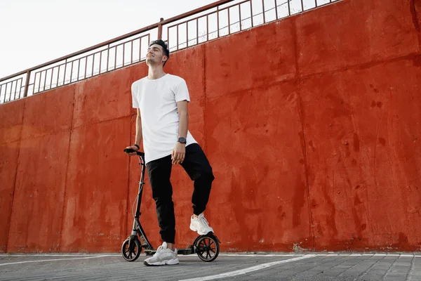 Jonge kerel gekleed in jeans en t-shirt staat met een scooter tegen een geschilderde betonnen muur op de zomerdag in de stad. — Stockfoto