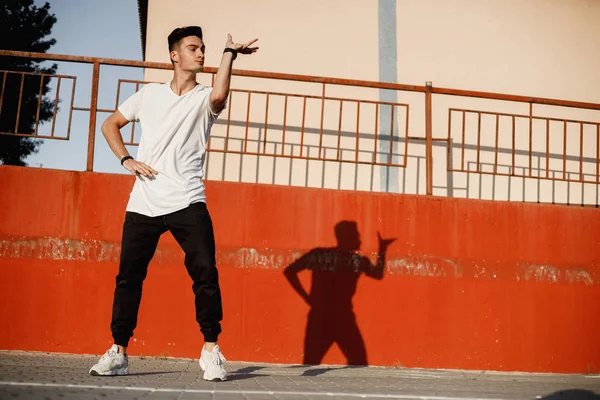 Elegante jovem vestido de jeans e t-shirt branca está dançando freio na rua contra uma parede de concreto pintado no dia ensolarado — Fotografia de Stock