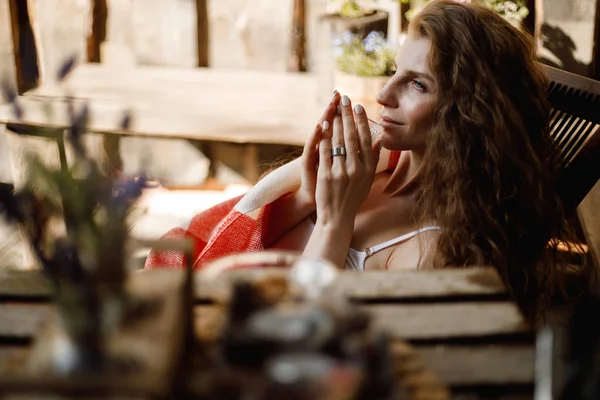 Jeune femme enveloppée dans un plaid rouge et blanc s'assoit dans une chaise pliante avec un verre d'eau à la main à côté d'un mur en bois — Photo